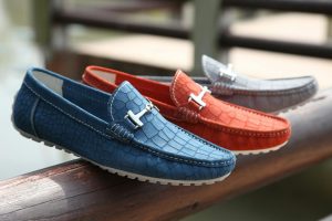Tre par loafers för herr i blått, orange och grått placerade på ett träräcke.
